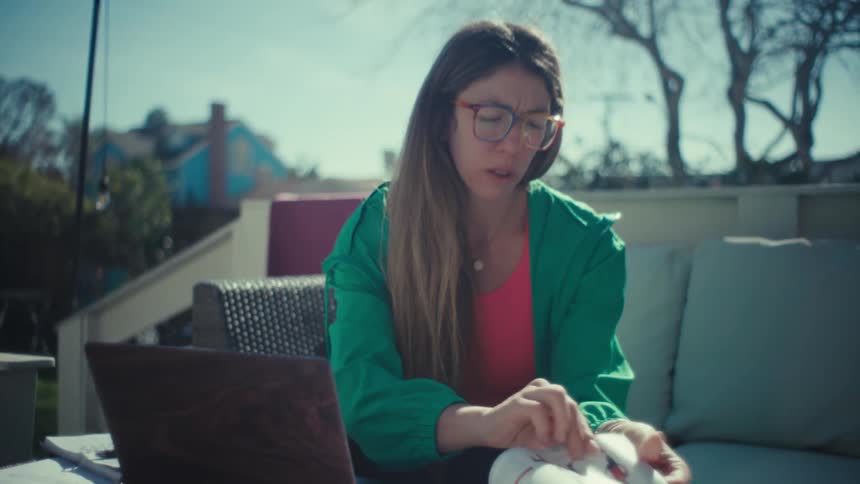 A woman wearing a green jacket and colorful glasses sits on an outdoor patio flipping through a textbook and typing on a laptop computer while studying in the bright sunlight