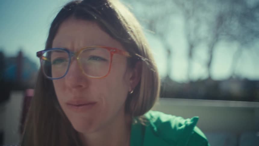 A woman wearing colorful glasses and a green jacket sits outdoors and looks at a screen with a thoughtful expression while resting her chin on her hand in the bright sunlight
