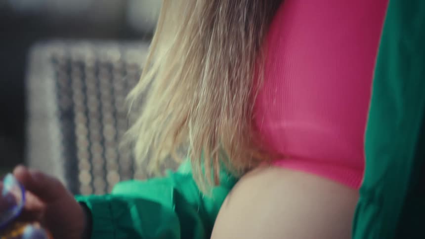 A pregnant woman wearing a green jacket and colorful glasses sits outdoors and types on a laptop computer before taking off her glasses and touching her stomach with a tired expression in the bright sunlight