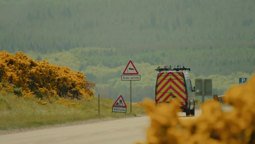 A yellow and red van drives down a paved road past a warning sign for side winds with a lush green forest in the background during the day