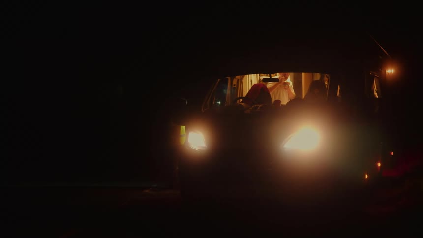 A person sits inside a van with the headlights on and an interior light glowing while parked in a dark area during the night
