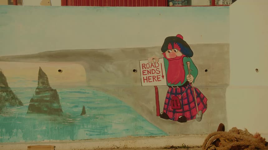 A mural on a wall depicts a cartoon character in a kilt holding a sign that says road ends here next to a painting of a coastline with large rock formations in the ocean during the day