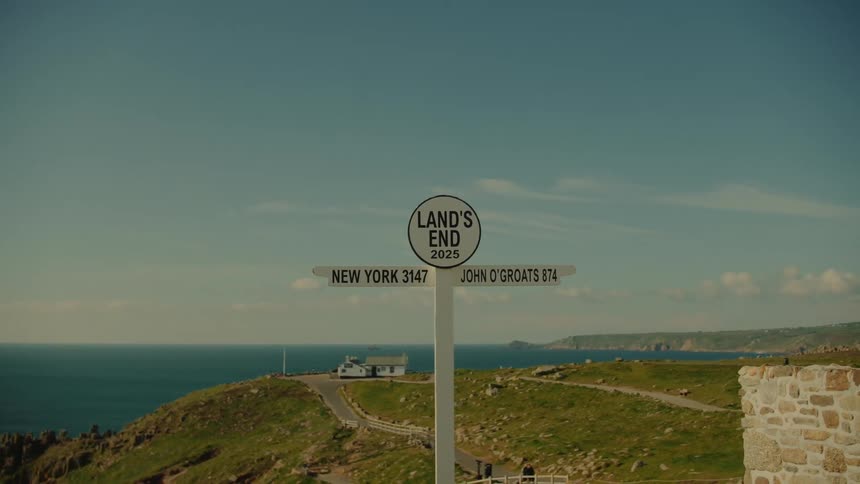 The camera zooms in on the famous lands end signpost against a scenic coastal backdrop