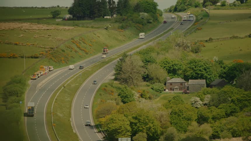 Winding highway cuts through a rural area with moving vehicles and houses among vegetation