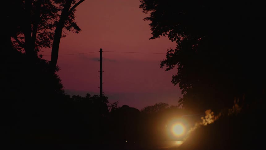 A car passes by on a shadowed road framed by trees with a dramatic red orange sunset visible in the distance