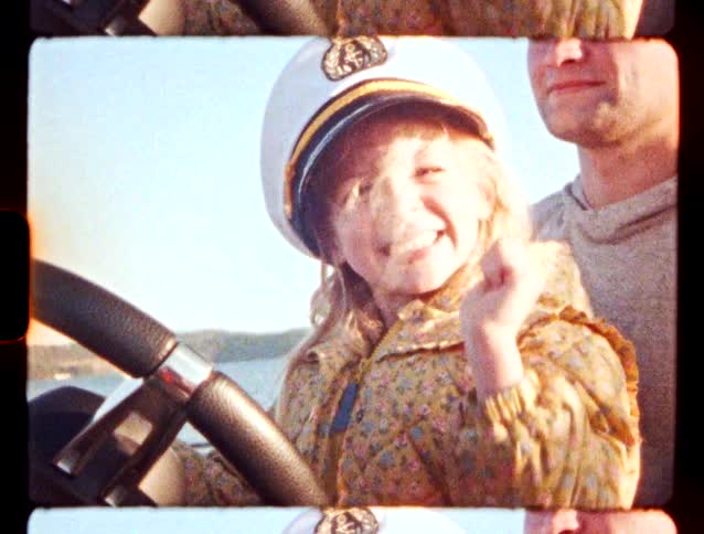 A young girl wearing a floral jacket and a white captains hat smiles while holding the steering wheel of a boat next to a man on a sunny day