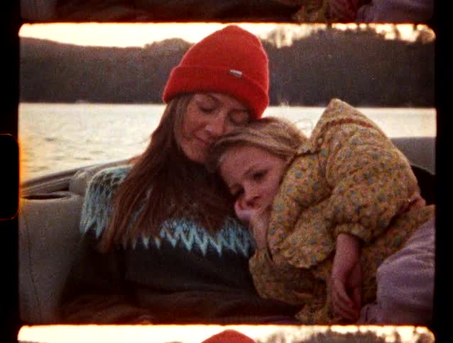 A woman in a red beanie gently holds a young child in a yellow patterned jacket as they sit together on a boat by the water