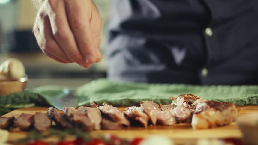 A chefs hand sprinkles salt onto sliced steak arranged on a wooden cutting board preparing it for serving