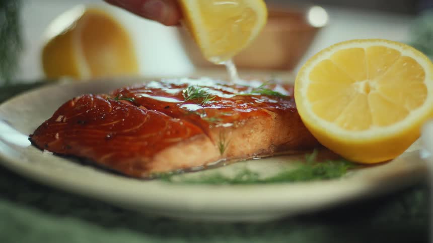A person squeezes juice from a lemon wedge onto a cooked salmon fillet garnished with dill on a plate during the day
