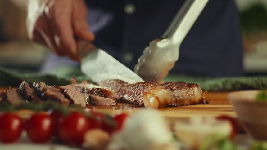 A chef uses a large knife and tongs to slice a cooked steak into thin strips on a wooden cutting board the foreground features a shallow focus on cherry tomatoes and other ingredients highlighting the freshly cut meat