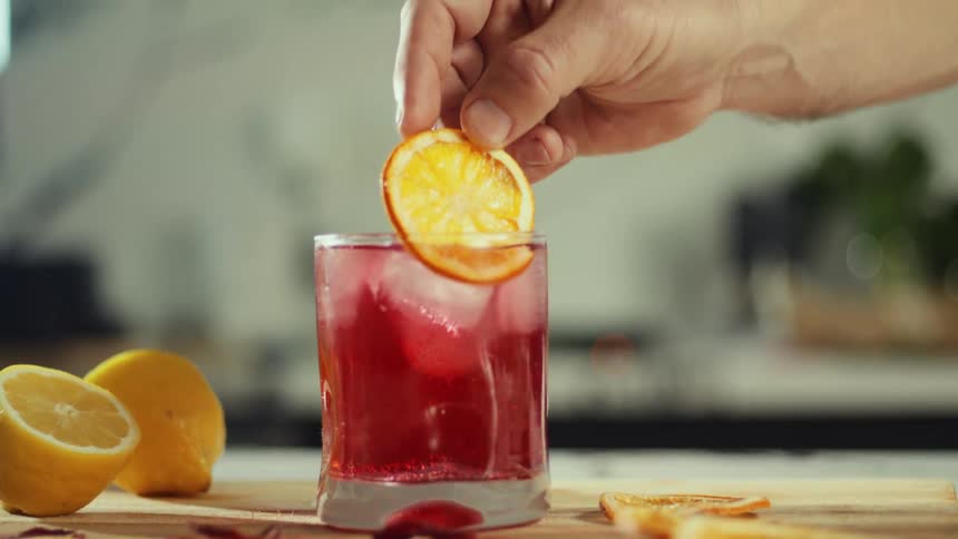 A hand places a dried orange slice onto the rim of a glass filled with a red iced beverage the drink sits on a wooden surface surrounded by fresh lemons and additional dried citrus garnishes