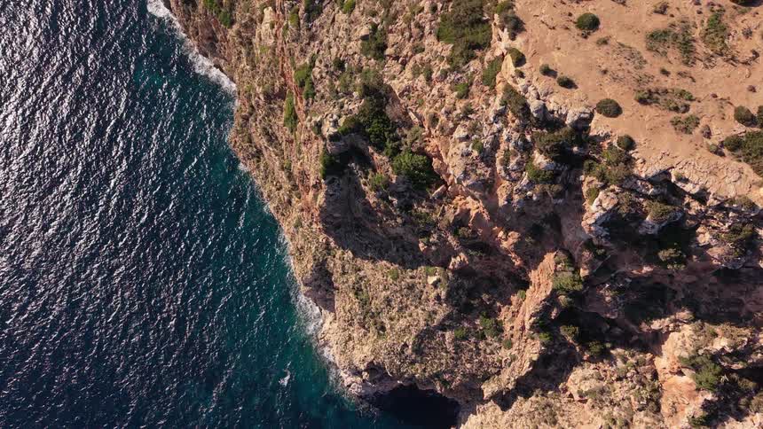 A top down drone shot captures the rugged edge of a steep rocky cliff meeting the deep blue ocean as small waves crash against the base and sunlight casts long shadows over the dry earthen ground and scattered green bushes above