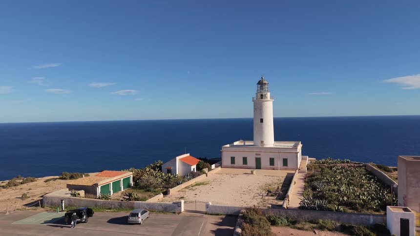 A wide aerial view shows a lighthouse and several nearby buildings at the end of a road along a rocky coastline overlooking the ocean