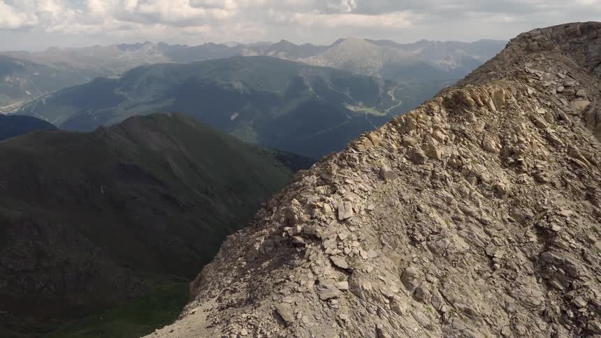 A wide aerial view captures a jagged rocky ridge in the foreground which slopes down toward a vast mountain range under a cloudy sky