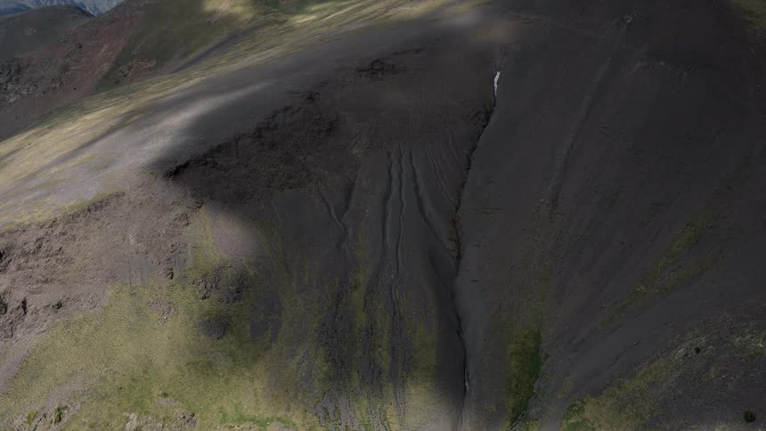 An aerial view shows a steep dark mountain slope cut by a narrow ravine with patches of green vegetation