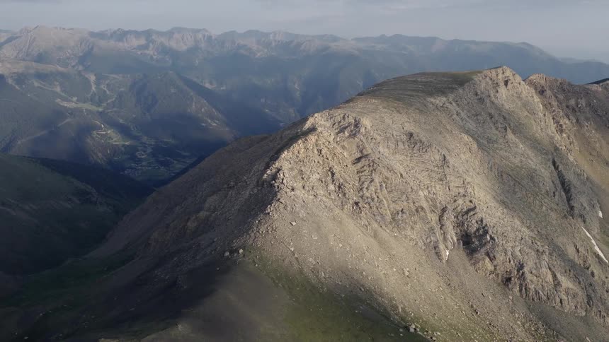 This aerial footage showcases a rugged mountain ridge under a clear sky highlighting the sharp contrast between the sunlit slopes and the deep shadows of the valley below the camera pans across the rocky terrain revealing distant peaks and a sprawling