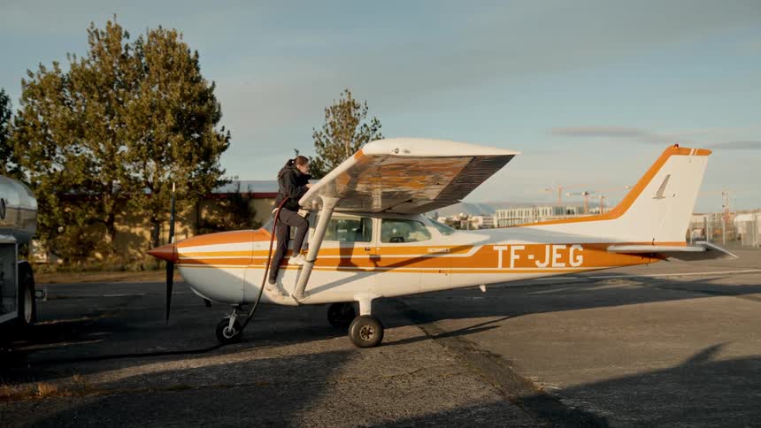 A person stands on a small orangeandwhite propeller airplane parked on the tarmac refueling it under the wing with trees and airport buildings visible in the background