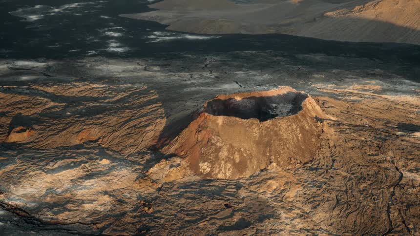 An aerial view pans right across a desolate volcanic crater landscape under the warm glow of late afternoon sun