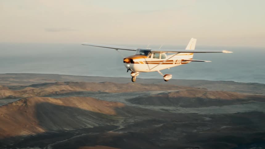 A small white and orange airplane flies over a sprawling city toward distant mountains under a pale sky during the late afternoon