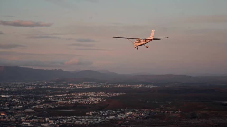 A small white and orange airplane flies over a sprawling city toward distant mountains under a pale sky during the late afternoon