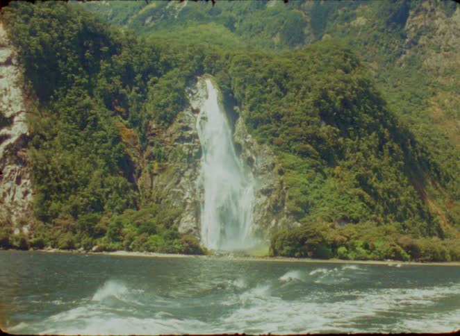 A scenic view of a large waterfall cascading down a lush green mountainside into a body of water the footage appears to be shot from a moving boat showing whitecapped waves in the foreground against the dramatic coastal landscape