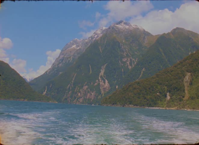 A scenic view shows a boats wake trailing across blue water toward a towering green mountain with snow near its peak under a partly cloudy sky