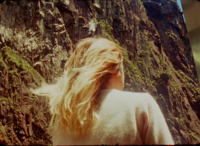 A young blonde woman standing beside a mountain rock with the wind blowing her hair at midday