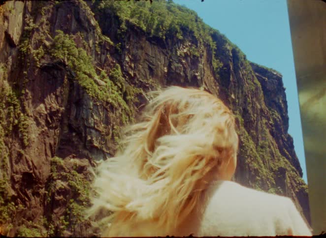 A woman with blonde hair stands with her back to the camera as wind blows through her hair in front of a massive steep rock cliff during the day