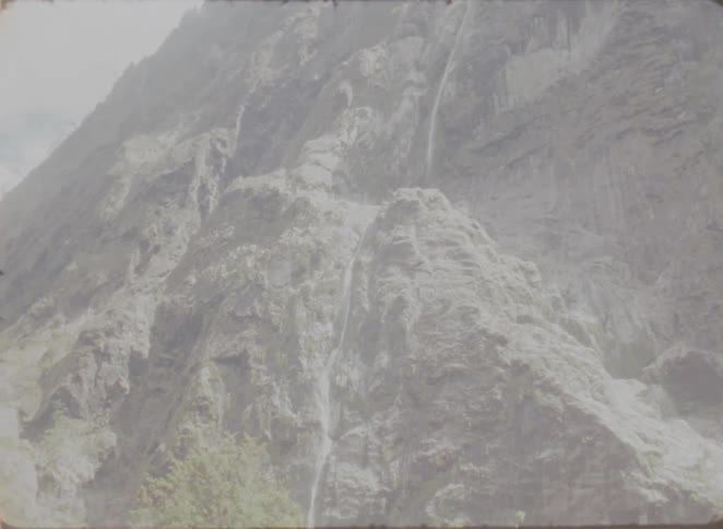 A view of a tall rocky mountain face with several thin waterfalls flowing down the steep cliffs toward green trees at the base on a bright day