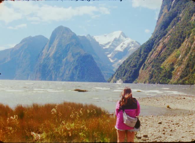 A woman with long brown hair wearing a pink jacket and a white shoulder bag stands on a rocky shore looking toward large green mountains and a snow peaked mountain under a blue sky