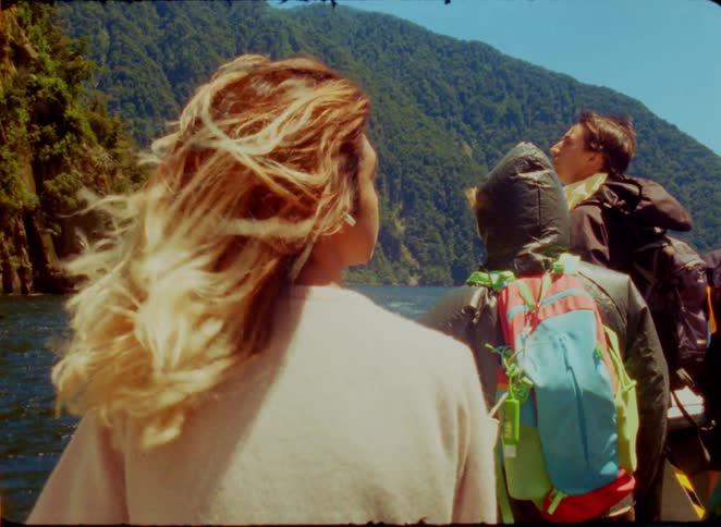 A group of friends stands on a boat looking toward a steep green mountain under a clear blue sky during the day