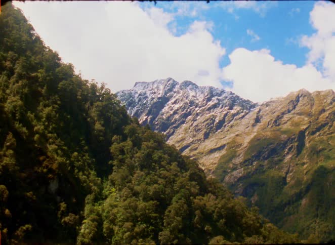 A scenic view of lush forested mountains beneath a bright blue sky filled with drifting white clouds a small airplane is visible flying high above the rugged snowdusted peaks as the camera pans across the landscape