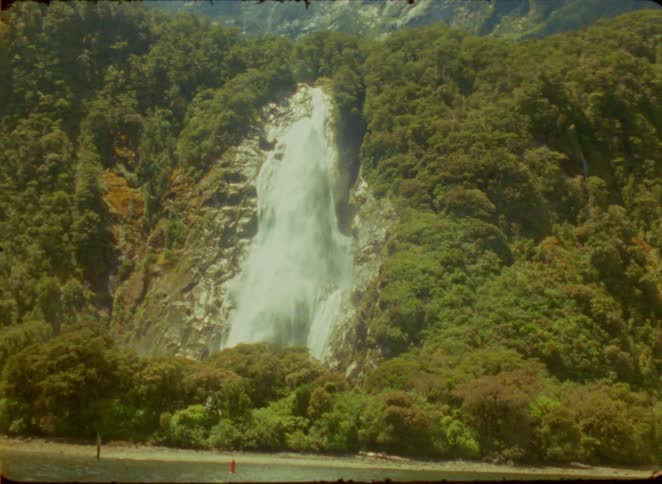 A powerful waterfall cascades down a lush green mountain slope into a valley surrounded by dense forest under a clear sky during the day