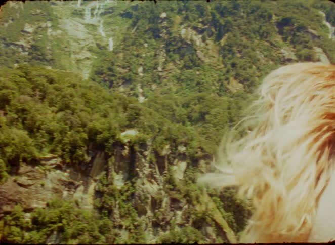 A young blonde woman on an airplane flying over a mountain covered in green vegetation with the wind blowing her long hair around at midday