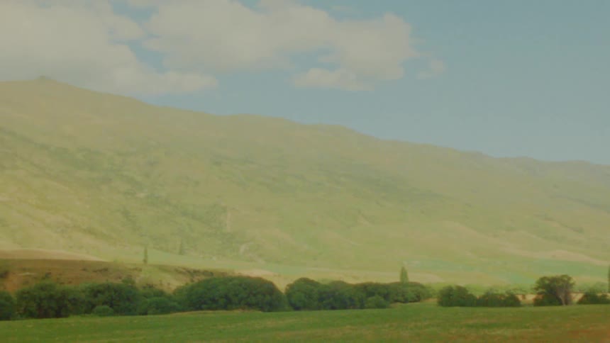 Vintage film footage shows a green valley and a field of tall grass with large brown mountains under a blue sky with soft white clouds