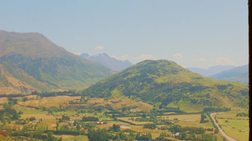 A scenic pan across a lush sundrenched valley nestled among rolling green hills and distant mountain peaks the landscape is dotted with patches of forest and rural settlements capturing a peaceful expansive natural vista