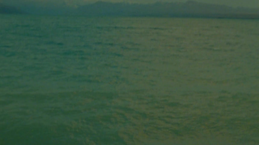 The view ascends from choppy green water to reveal mountains under a cloudy sky
