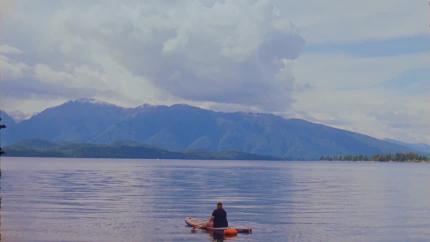 A person sits on a paddleboard in the middle of a calm expansive lake surrounded by distant mountains the footage uses a vintage film style with a soft color palette and grainy texture