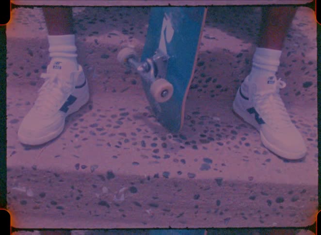 A person wearing white new balance sneakers and high socks sits on concrete steps while holding a blue skateboard the footage features a vintage grainy film aesthetic with a distinct purple and pink color grade