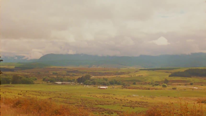 A wide green valley with scattered buildings sits beneath a cloudy sky and distant mountains during the day