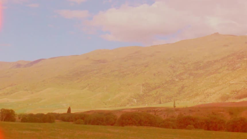 Rolling green hills and distant mountains sit under a blue sky with soft white clouds before a bright yellow light leak covers the entire frame