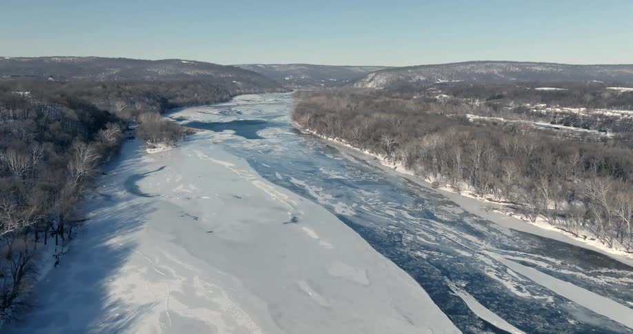 Partially frozen river runs through a valley surrounded by hills and trees in winter