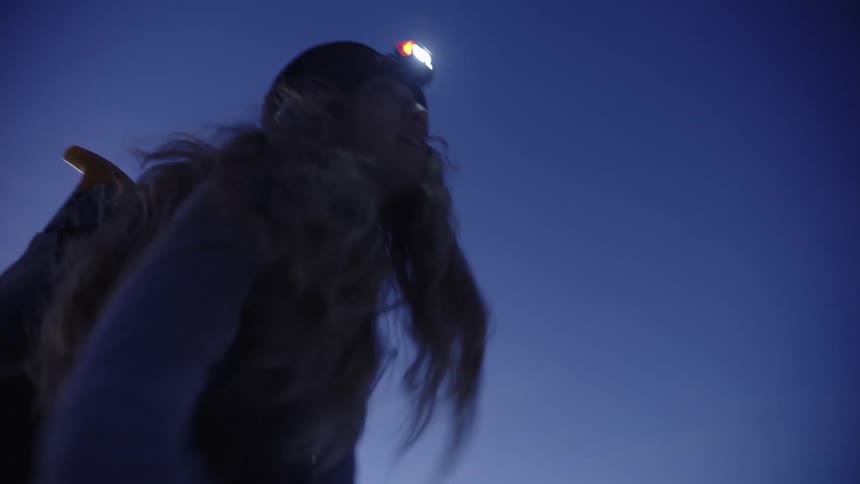A young woman climbing a snowy mountain in the evening wearing skiing equipment and a headlamp holding ski poles as she ascends