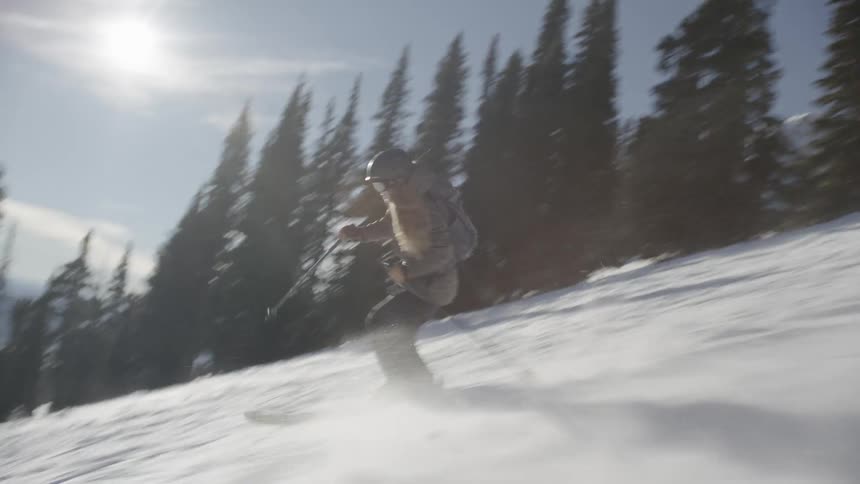 A female skier wearing a grey jacket and helmet carves through deep powder on a steep mountain slope lined with tall evergreen trees under a bright sun