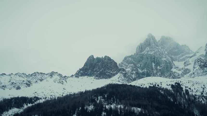 Forested mountain with snow and ice covering the rough terrain of mountains with peaks high in the cloudy winter sky