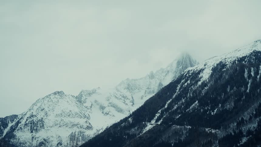 A view of tall mountains covered in snow and dark green trees under a grey and cloudy sky