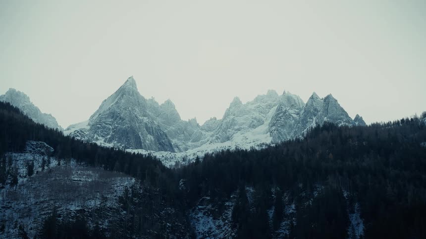 A series of sharp jagged mountain peaks covered in snow tower over a dense dark forest under a stark pale sky
