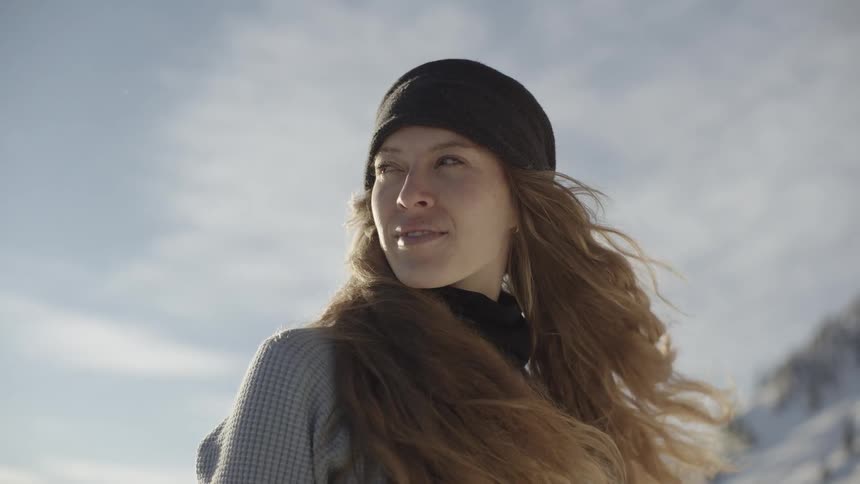 A young woman with long wavy hair stands in a snowy mountain landscape wearing a black beanie and sweater