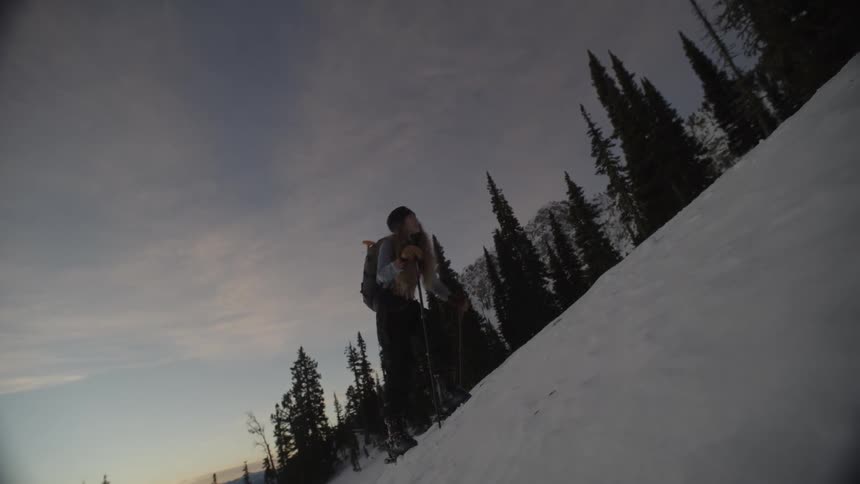 A man and a woman stand on a snowy mountain slope at dusk and use their ski poles to walk uphill past tall dark trees
