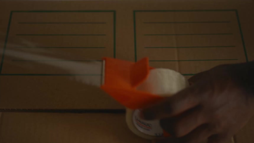 Hand using orange tape dispenser to seal cardboard box with green labels